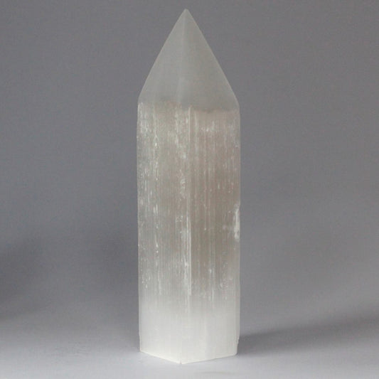 A selenite crystal point stands upright on a pale gray surface, its translucent, vertically striated form tapering to a peak at the top.