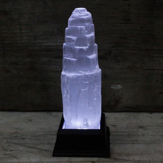 A selenite crystal tower glows with blue light, standing on a black base. The tower is lit from underneath. It sits on a wooden surface against a backdrop of weathered wood.