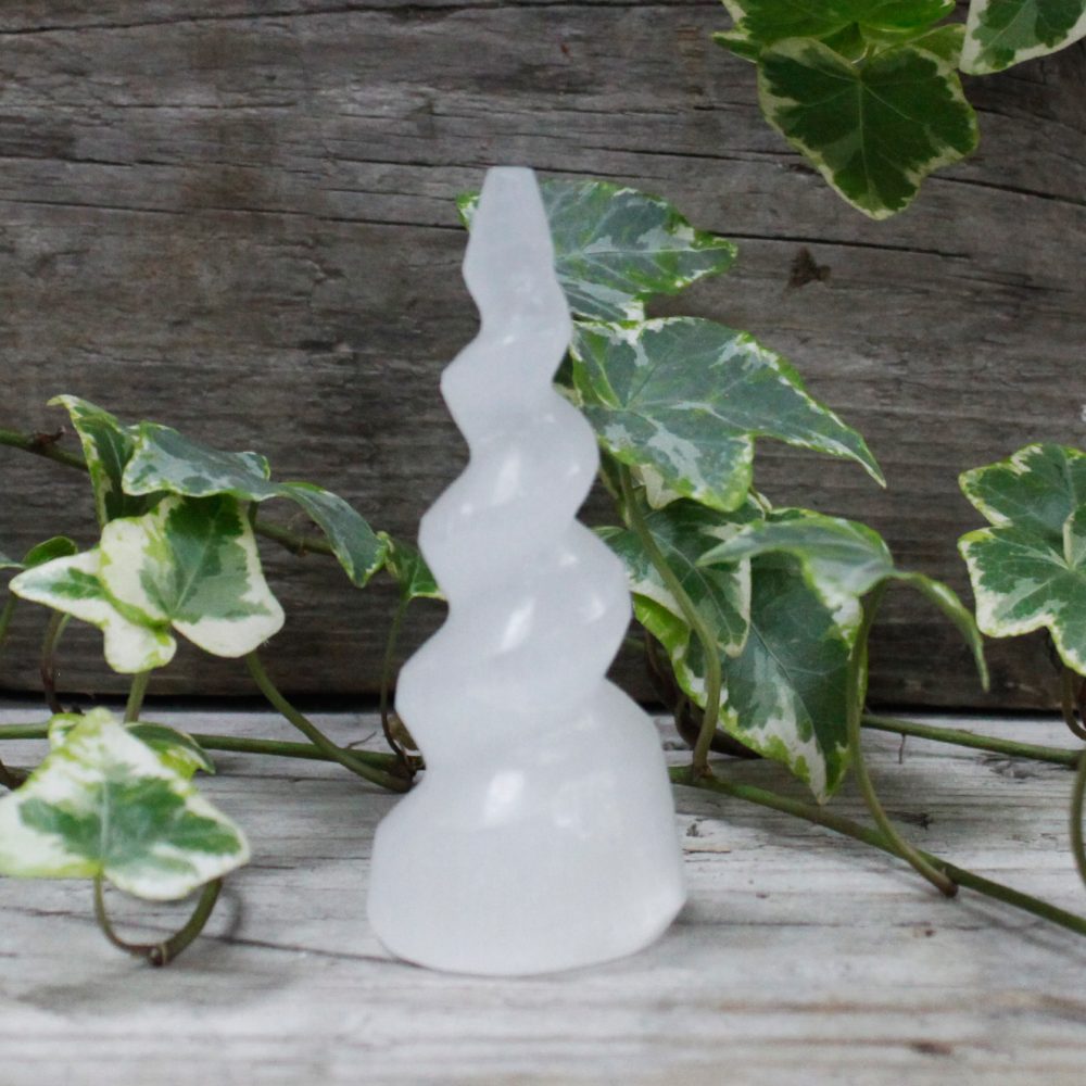 A white selenite tower with a spiral shape stands on a wooden surface. Green and white ivy surrounds the base and background.