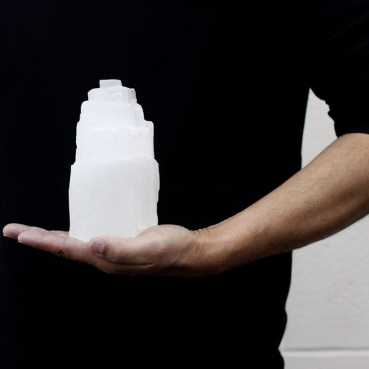 A hand holds a tiered, opaque white crystal block, resembling a miniature tower. The person, dressed in black, stands against a pale gray background.