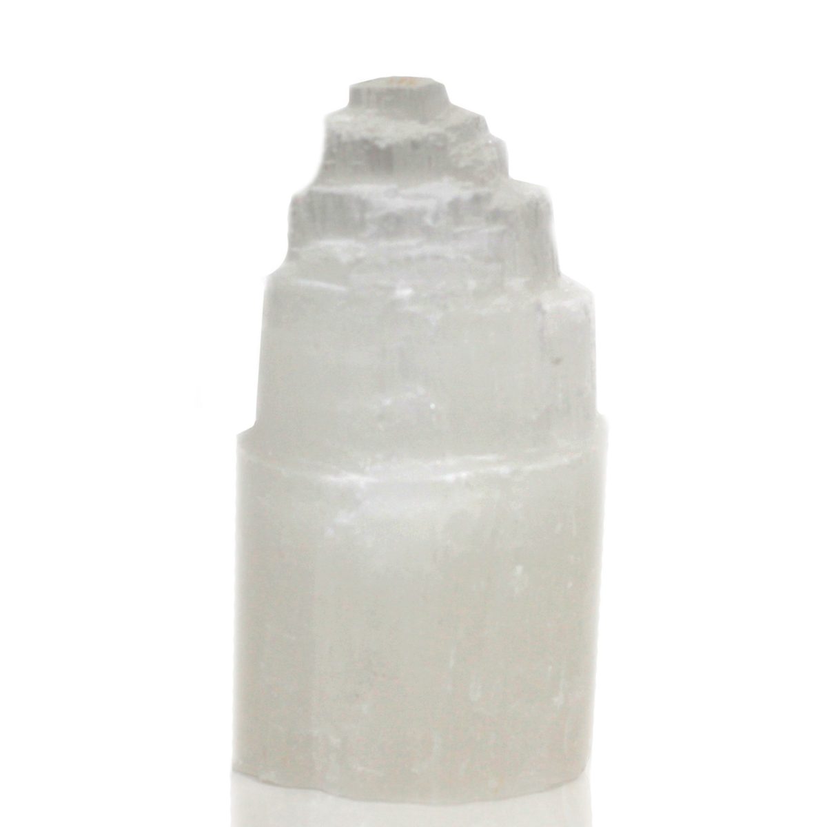 A tower-shaped selenite crystal point is set on a glossy white surface. It features a tiered, cylindrical body that tapers into a multi-layered, stepped peak.