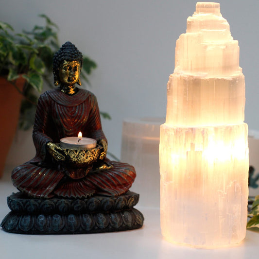 Buddha statue sits in meditation, holding a lit candle. A glowing selenite lamp stands to the right, on a white surface with plants in the background.