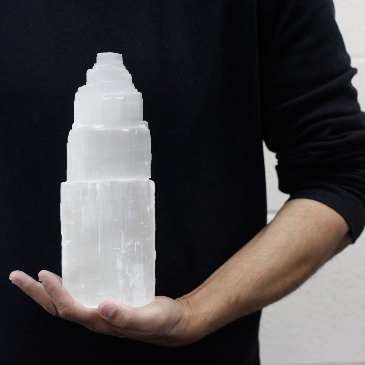 A man in a black sweatshirt holds a tiered, white selenite tower in his hand. The background is a plain, light-colored wall.