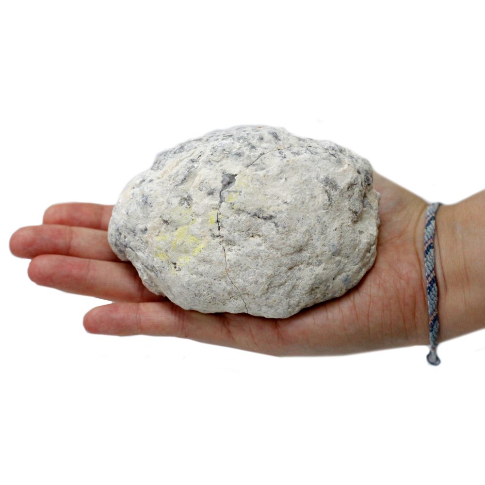 A large, rough, off-white rock with yellow inclusions rests in the palm of a hand. The hand wears a multi-colored braided bracelet, against a white background.