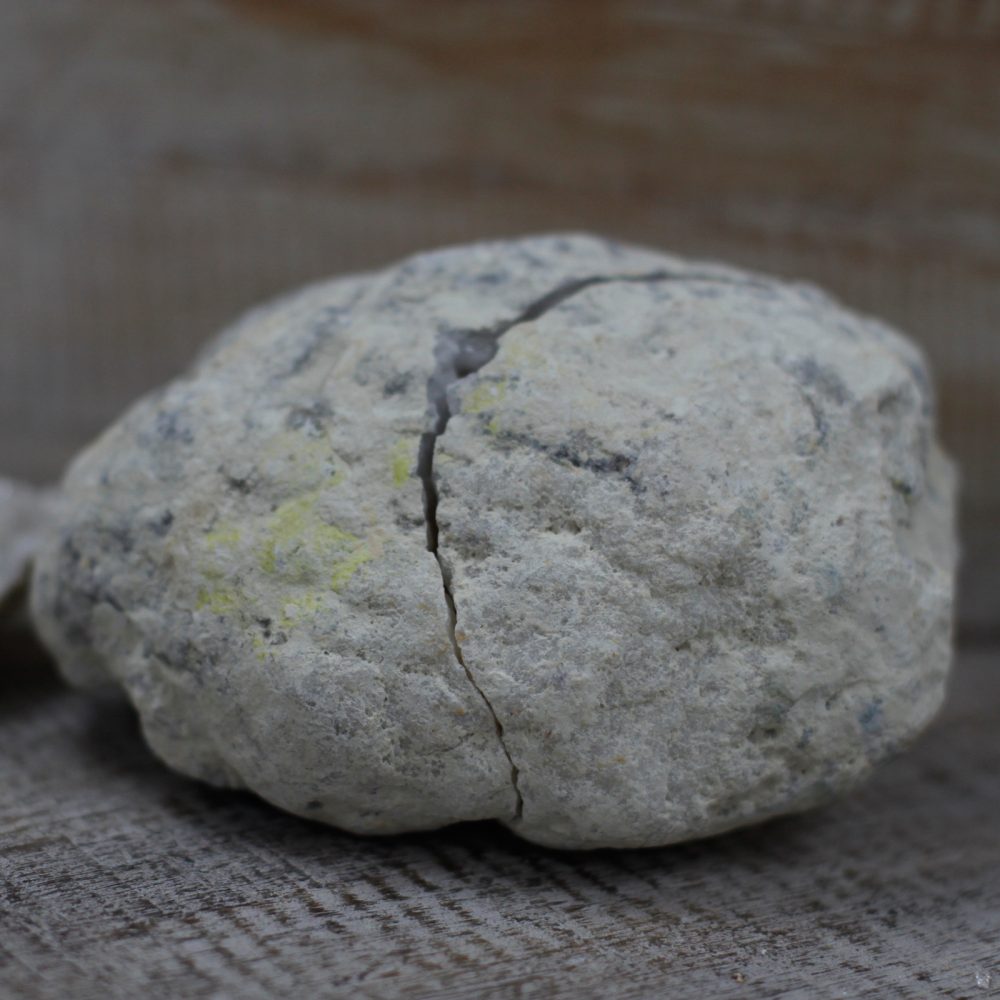 A cracked, roughly spherical, gray rock rests on a light wooden surface. A thin seam of white crystals is exposed by the crack.