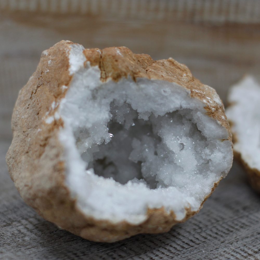 A geode, split open, displays white crystal formations inside its light-brown, rock-like exterior. It sits on a light-brown, textured surface.