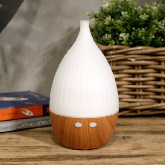 The white, bulbous humidifier sits on a wooden surface. Behind it are books, a wicker basket, and greenery, set against a neutral gray wall. The visible books read, "MARTEL," and "FLEMING CASINO ROYALE."