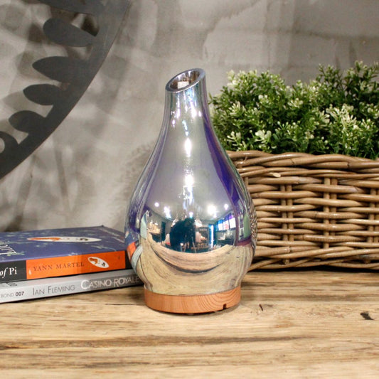 A reflective silver diffuser sits on a wooden table near books and a basket with greenery. The books are titled “Life of Pi” by Yann Martel and "Casino Royale" by Ian Fleming.