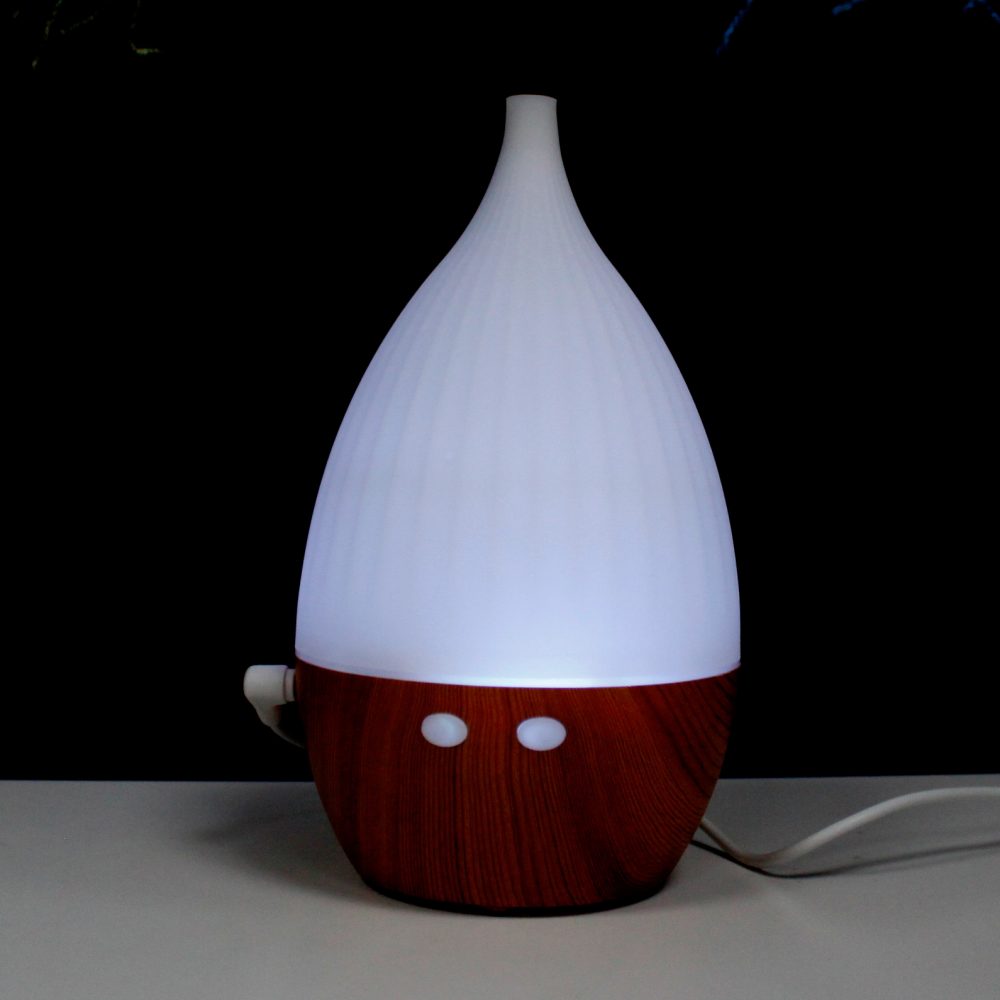 A teardrop-shaped aroma diffuser glows white from its top half while sitting on a white surface, its lower half appearing like reddish wood. A black background is present.