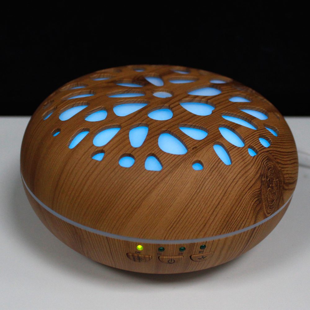 A wood-grain patterned diffuser sits on a white surface, glowing with blue light through its decorative perforations. The buttons at the base read "ON," "1H," "3H," and "6H."