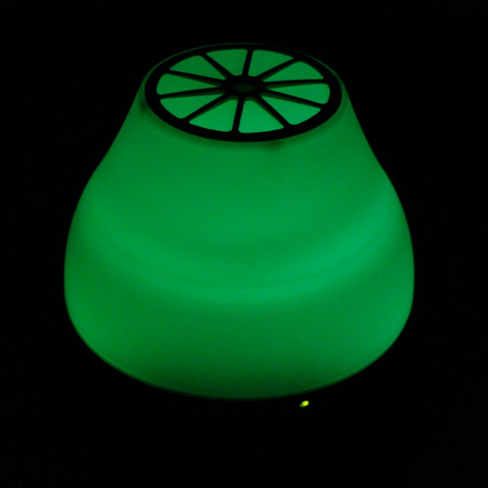 A glowing green pear-shaped lamp shines brightly in the dark. The top of the lamp has a black, circular, vented top.