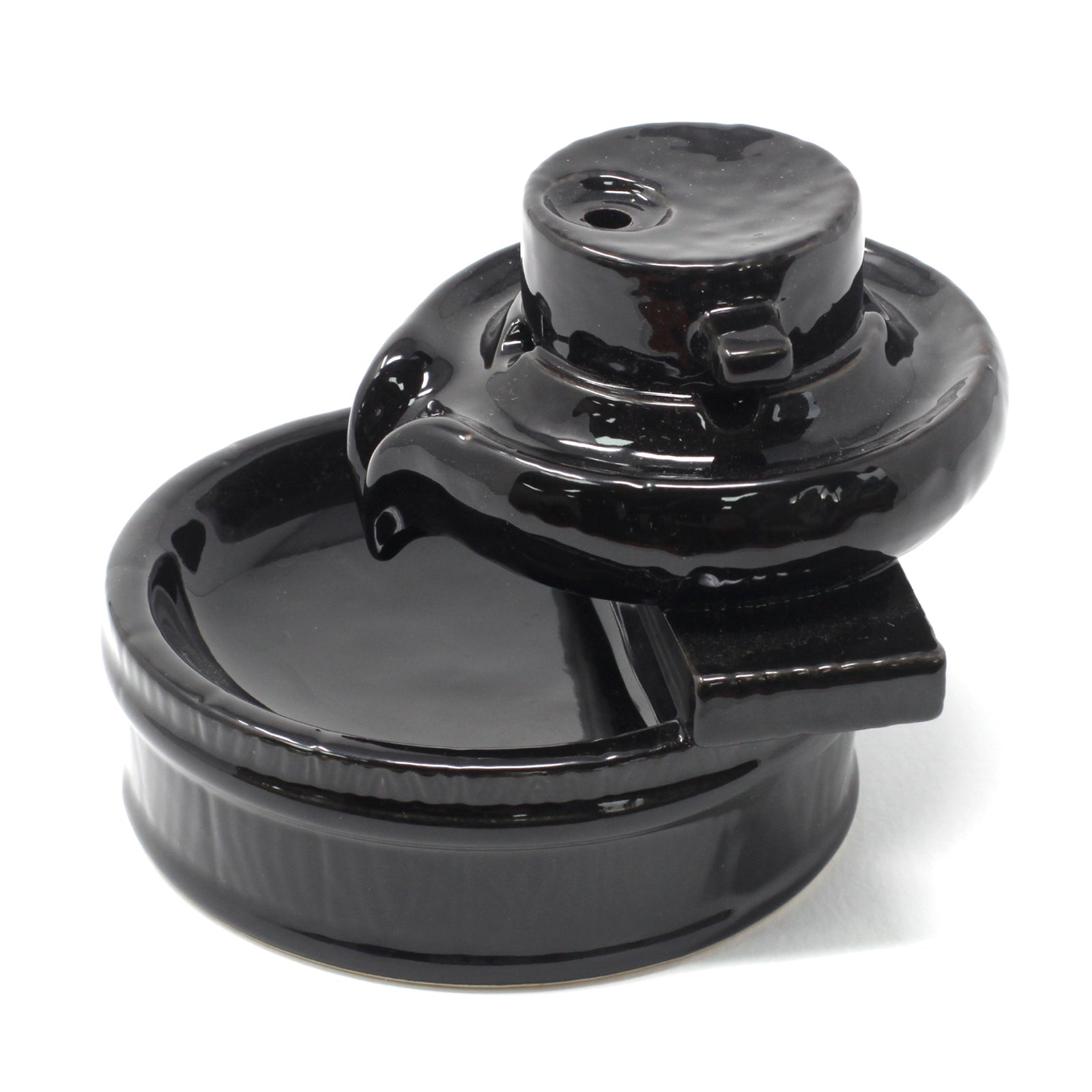 A glossy black ceramic fountainhead rests atop a shallow, circular basin, with a cylindrical structure on one side. The background is a plain white surface. No text appears.