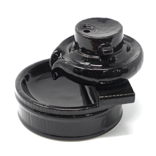 A glossy black ceramic fountainhead rests atop a shallow, circular basin, with a cylindrical structure on one side. The background is a plain white surface. No text appears.