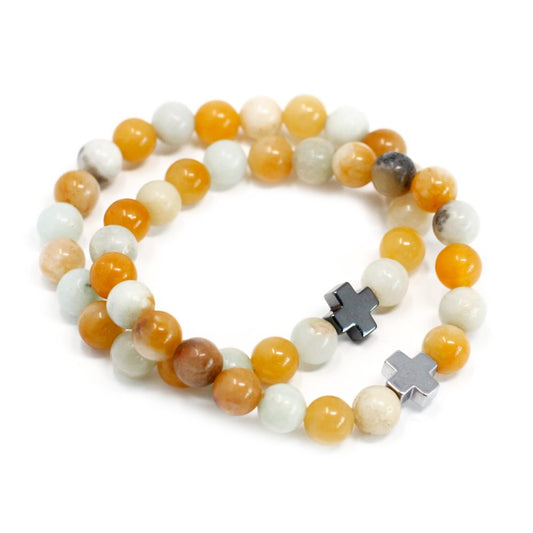 Two bracelets of variegated orange and white beads, each featuring a small, metallic cross charm, rest on a plain white surface.