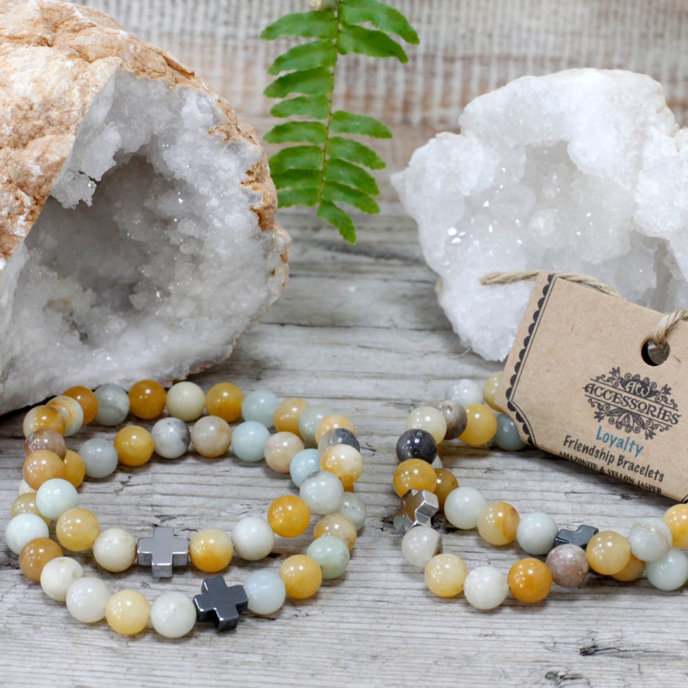 Two bead bracelets rest on a wooden surface, adorned with metal crosses. A tag reads "ACCESSORIES Loyalty Friendship Bracelets AMAZONITE & YELLOW JASPER". Crystals and a fern leaf decorate the backdrop.