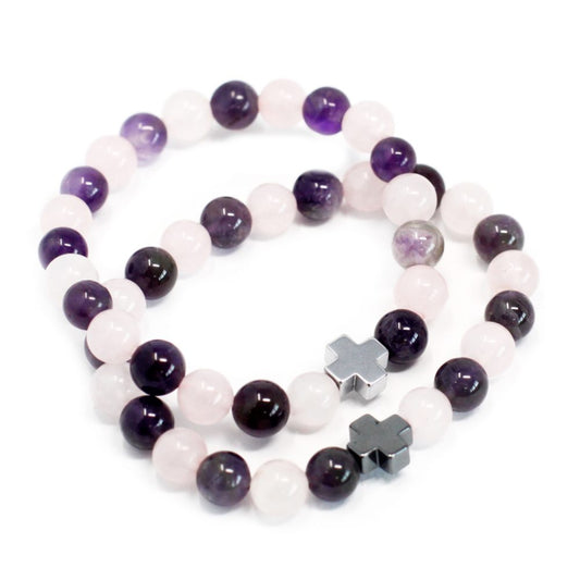 Two beaded bracelets rest on a white surface. The bracelets alternate between pale pink and dark purple beads and include small metal cross charms.
