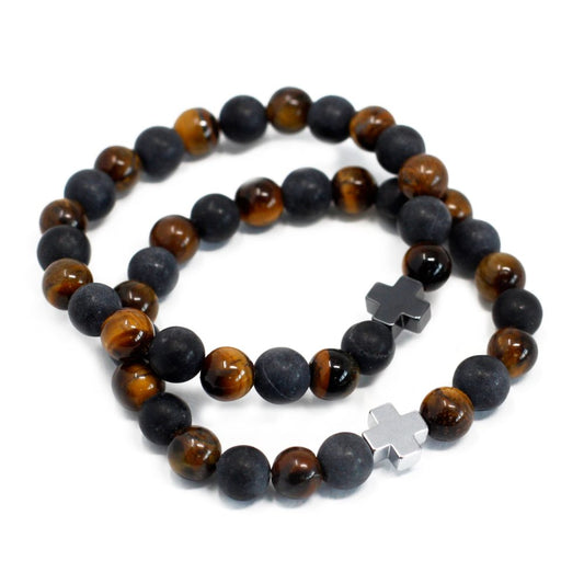 Two beaded bracelets lay intertwined, each alternating black and tiger-eye stones. One bracelet features a dark metallic cross, while the other has a silver one. They are on a plain white background.