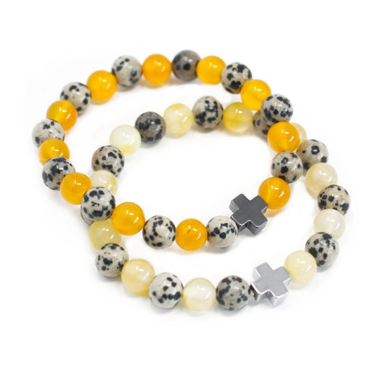 Two beaded bracelets are coiled, featuring orange, dalmatian-spotted, and pale yellow beads, each with a cross-shaped bead, set on a bright white background.