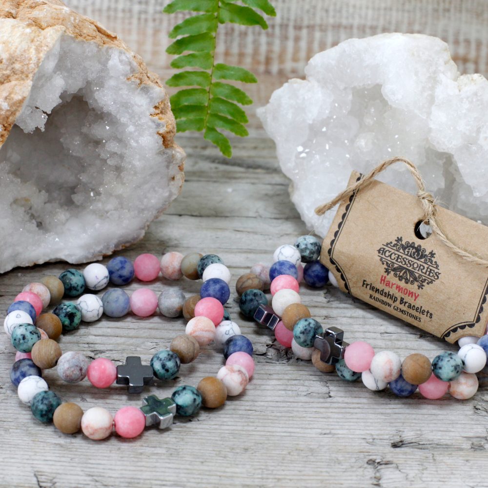 Two colorful beaded bracelets sit on a wood surface next to crystals, ferns, and a tag which reads “Accessories Harmony Friendship Bracelets Rainbow Gemstones.”