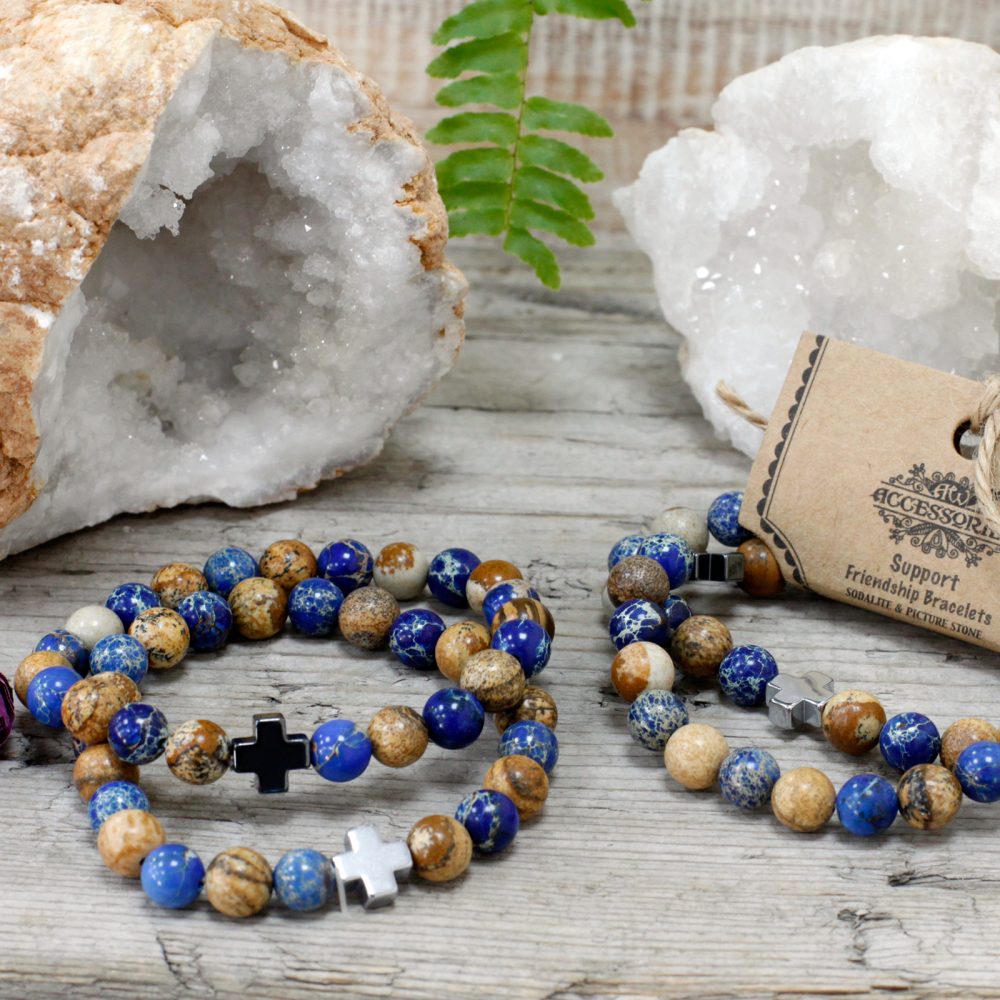 Two beaded bracelets, with cross charms, rest on a weathered wooden surface with two geodes and a sprig of fern.
The tag reads “Accessories Support Friendship Bracelets SODALITE & PICTURE STONE”.