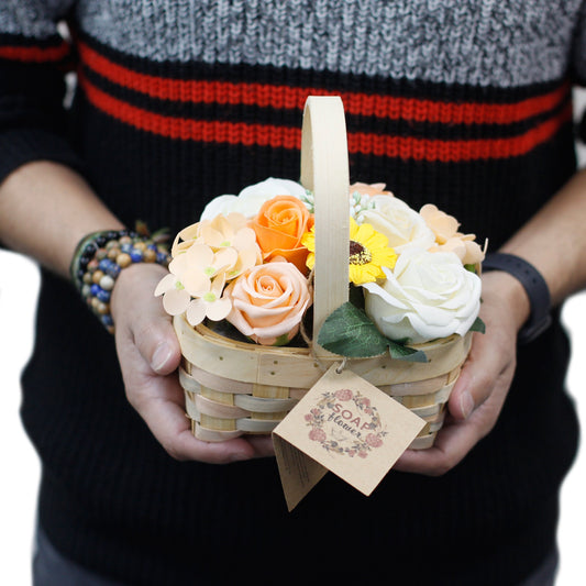 Small woven basket of soap flowers held by two hands, showcasing peach and white rose-shaped soaps and a yellow sunflower; person wears black and gray sweater with red stripes and beaded bracelets; tag reads SOAP flower