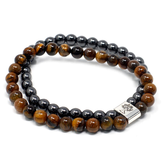 Two beaded bracelets, one of tiger's eye stones and one of grey hematite stones, are coiled in a stack on a white surface. The lower bracelet has a silver tree-of-life charm.