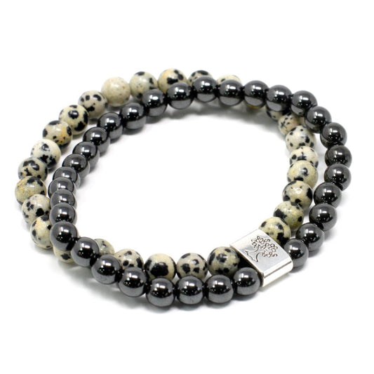 Two beaded bracelets, one of hematite beads, and one of dalmatian jasper, sit layered atop each other against a stark white background. One band has a tree-of-life charm.