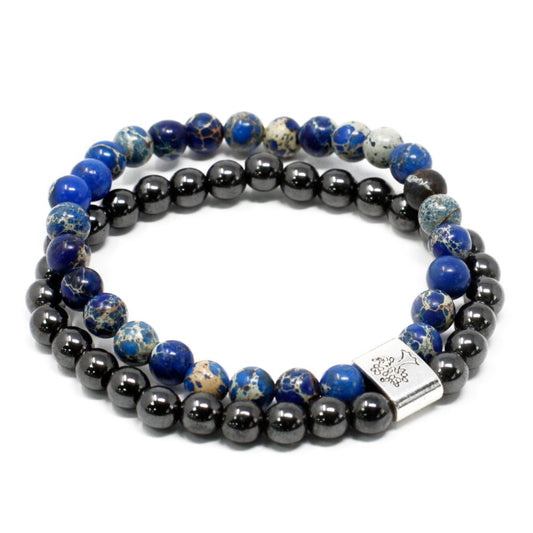 Two beaded bracelets are stacked on a white surface. One bracelet has blue-and-beige speckled beads, and the other has shiny, small, black beads with a silver charm.