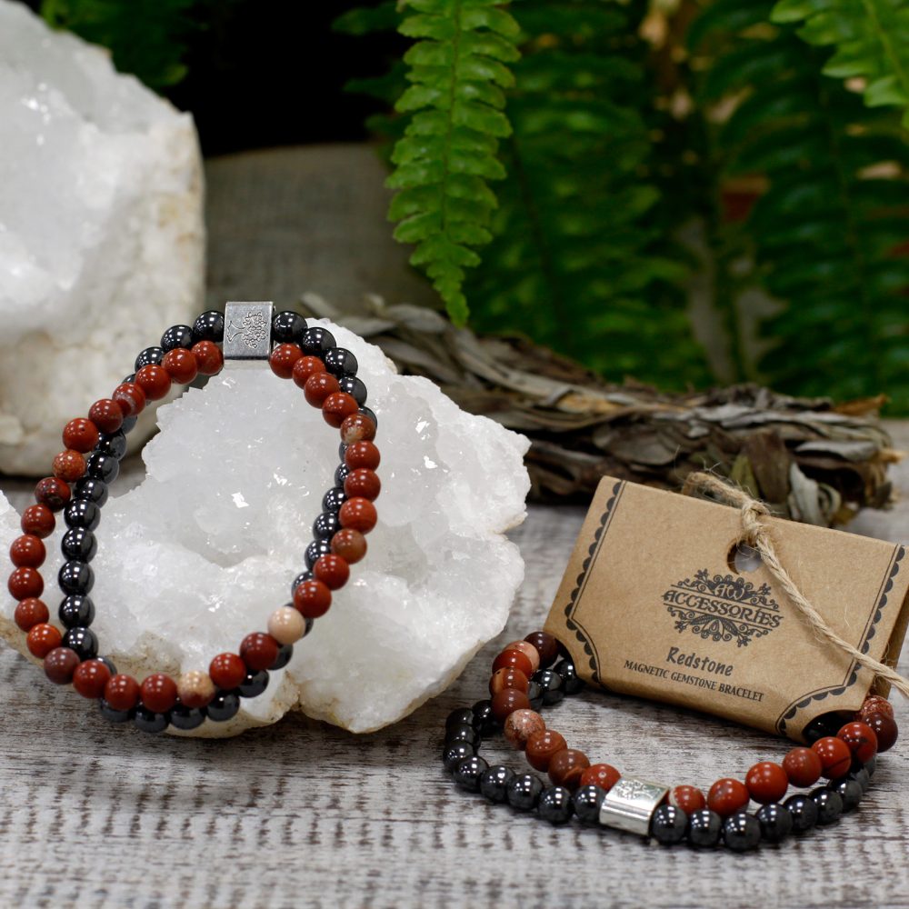 Two Redstone magnetic gemstone bracelets sit on a weathered wooden surface near a quartz cluster. A tag reads: “ACCESSORIES; Redstone; MAGNETIC GEMSTONE BRACELET." Greenery and a sage bundle are in the background.