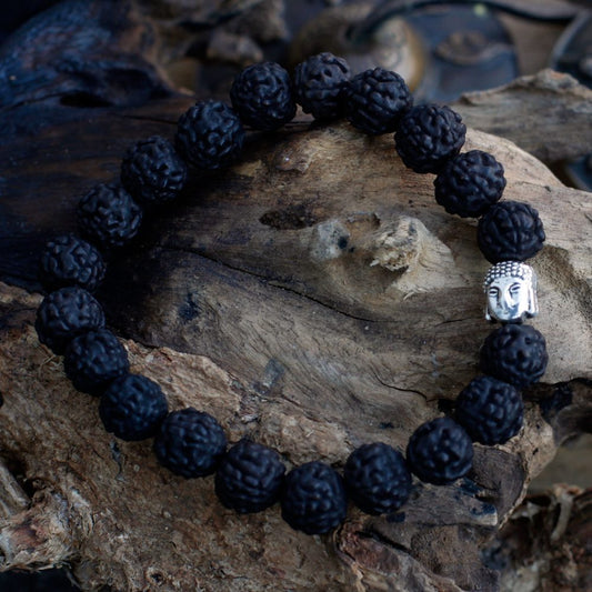 A beaded bracelet, with a silver Buddha face charm, rests on a gnarled piece of driftwood. The bracelet's beads are dark and textured.