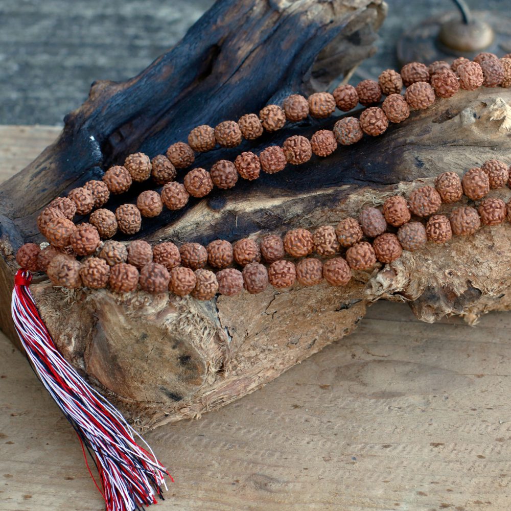 A mala, or beaded necklace, rests atop a piece of driftwood on a textured wooden surface. The necklace has a tassel with red, white, and black threads.