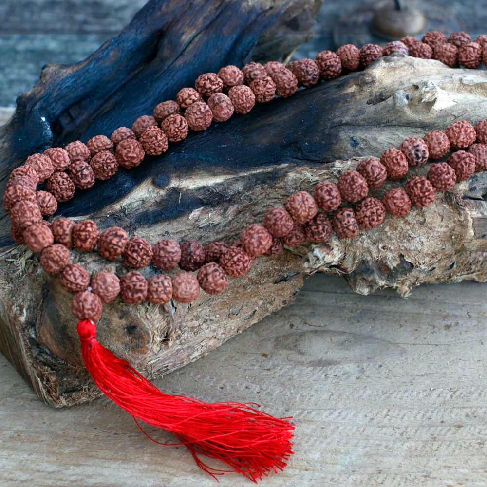 A strand of rudraksha beads drapes across a piece of rough wood. A red tassel hangs at one end, all resting on a light wood surface.