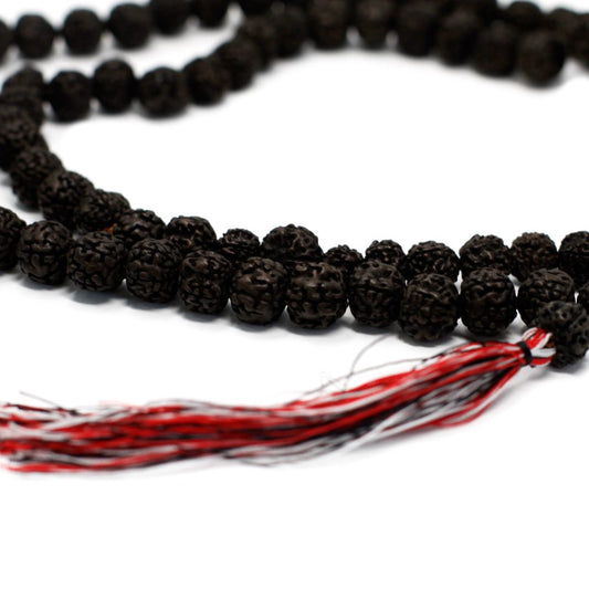 A mala rests on a white surface; the dark, round beads are strung together, with a red, white, and gray tassel dangling from one end.