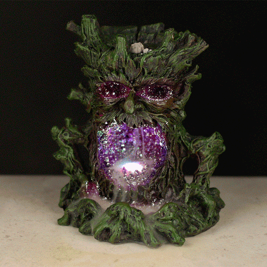 A tree-like incense holder emits smoke from a gaping mouth, surrounded by green, wood-textured features and set in a dimly lit environment. Light reflects off shiny purple accents inside.