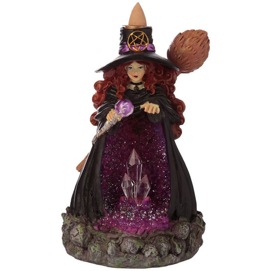 A witch figurine, holding a crystal ball-topped staff, stands on a mossy base with purple glitter and crystals. She wears a black hat with a pentacle and has red hair.