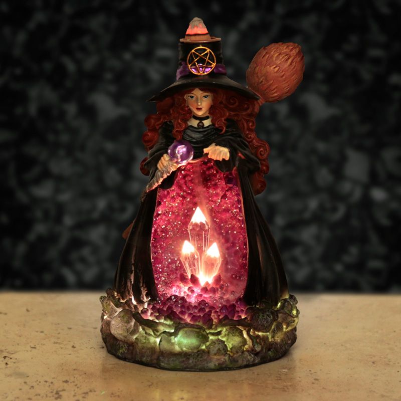 A statuette of a witch holding a crystal orb; her hat has a pentacle, a broom is on her back. A glowing crystal formation is displayed in front, over a rocky base.