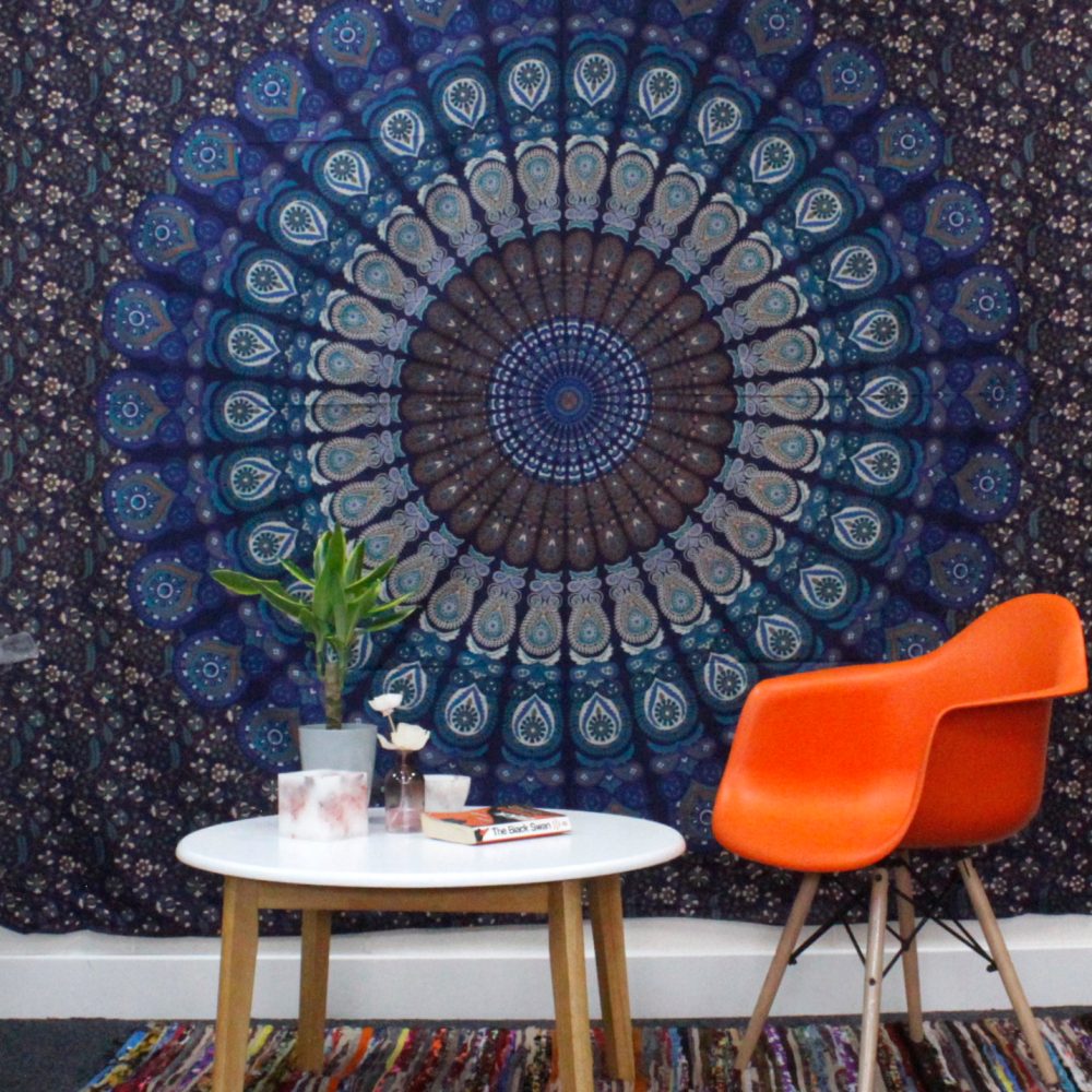 A mandala tapestry hangs on a wall; an orange chair and small table sit in front, holding plants and a book titled "The Black Swan".
