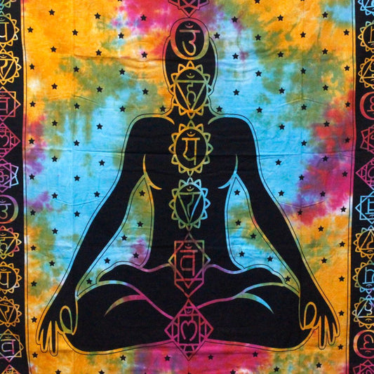 A black figure sits in a lotus position, displaying seven chakras, on a tie-dye tapestry with stars.