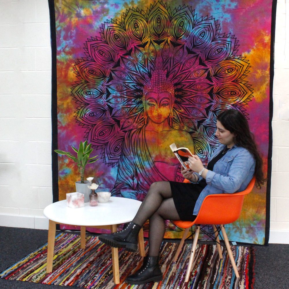 A woman sits on an orange chair, reading a book in front of a tie-dye tapestry depicting a Buddha. A small table with decorative objects is nearby.
