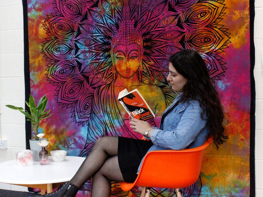 A woman sits in an orange chair, reading "Black Swan" in front of a tie-dye tapestry depicting Buddha.