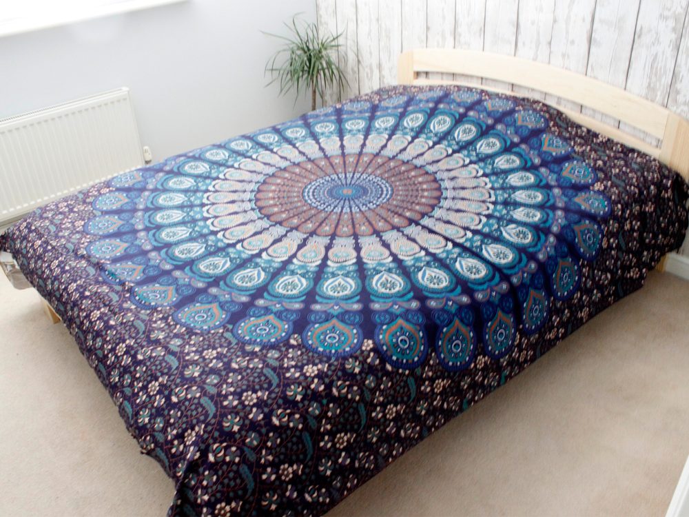 A mandala tapestry covers a bed, displaying teal, brown, and white patterns. It sits in a simply decorated bedroom.