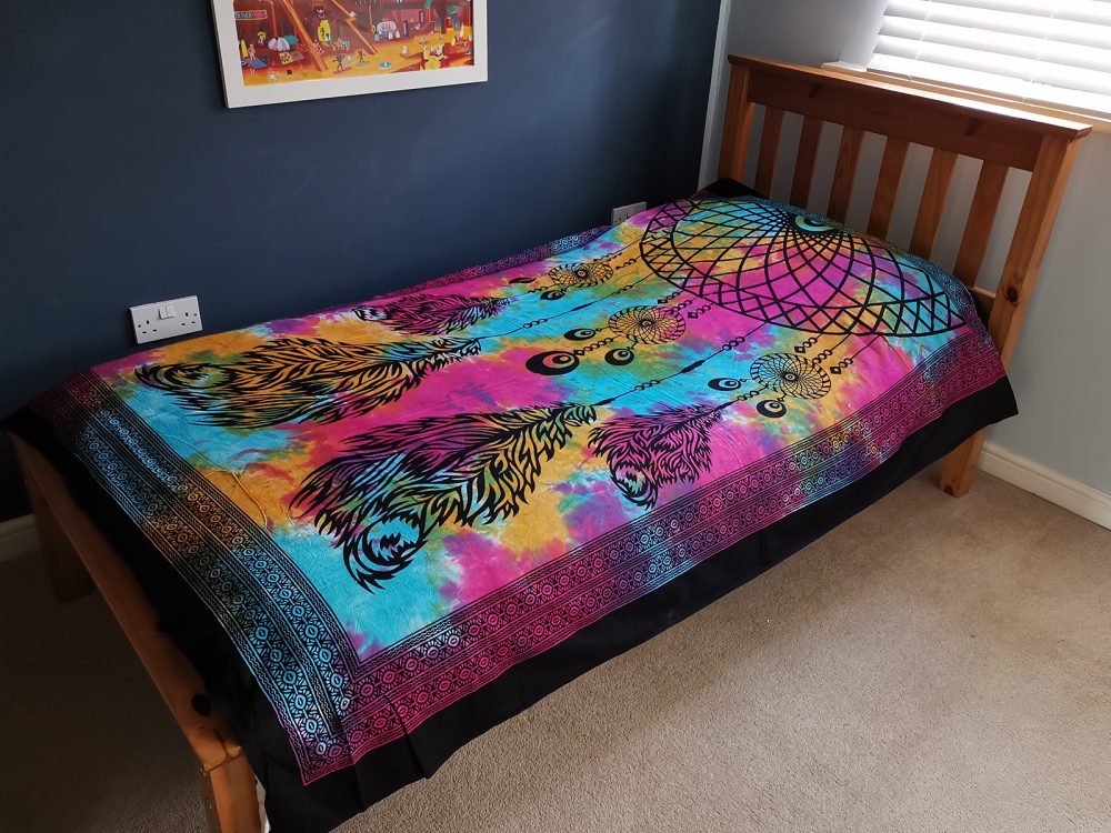 A tie-dye tapestry, featuring a dreamcatcher design with feathers, rests on a single bed in a child's bedroom.