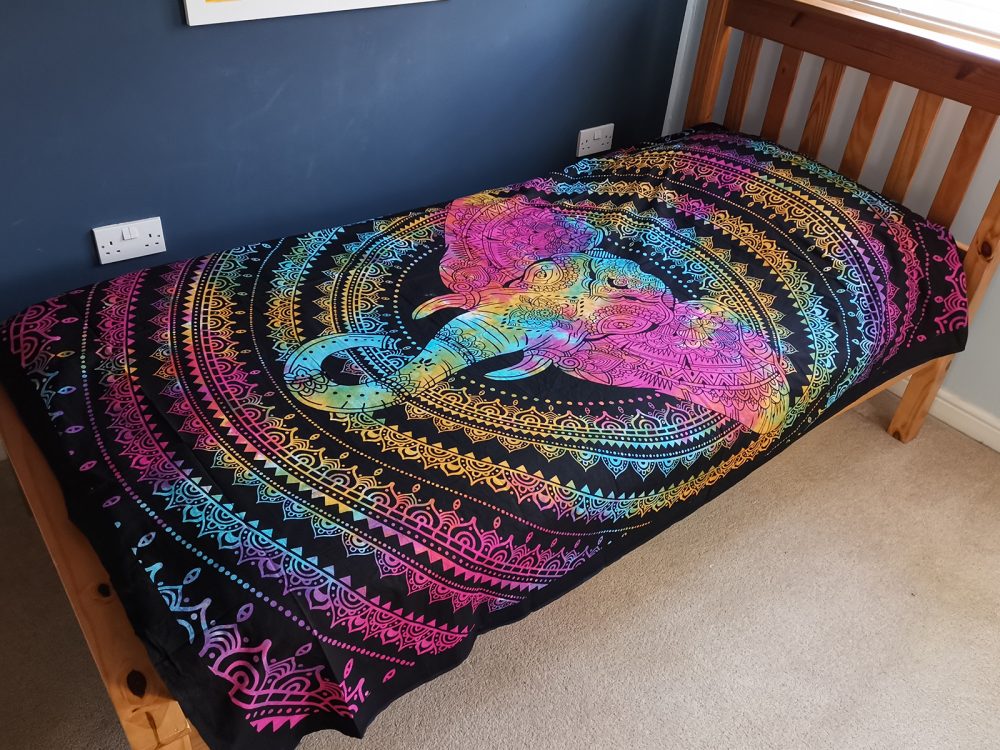 A tapestry depicting a rainbow-colored elephant mandala lies on a wooden bed frame in a bedroom.
