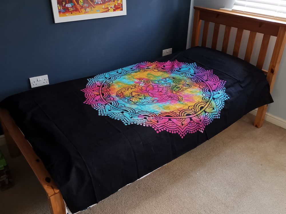 A tie-dye tapestry, featuring a Hamsa hand in a mandala, rests on a single bed in a child's bedroom.