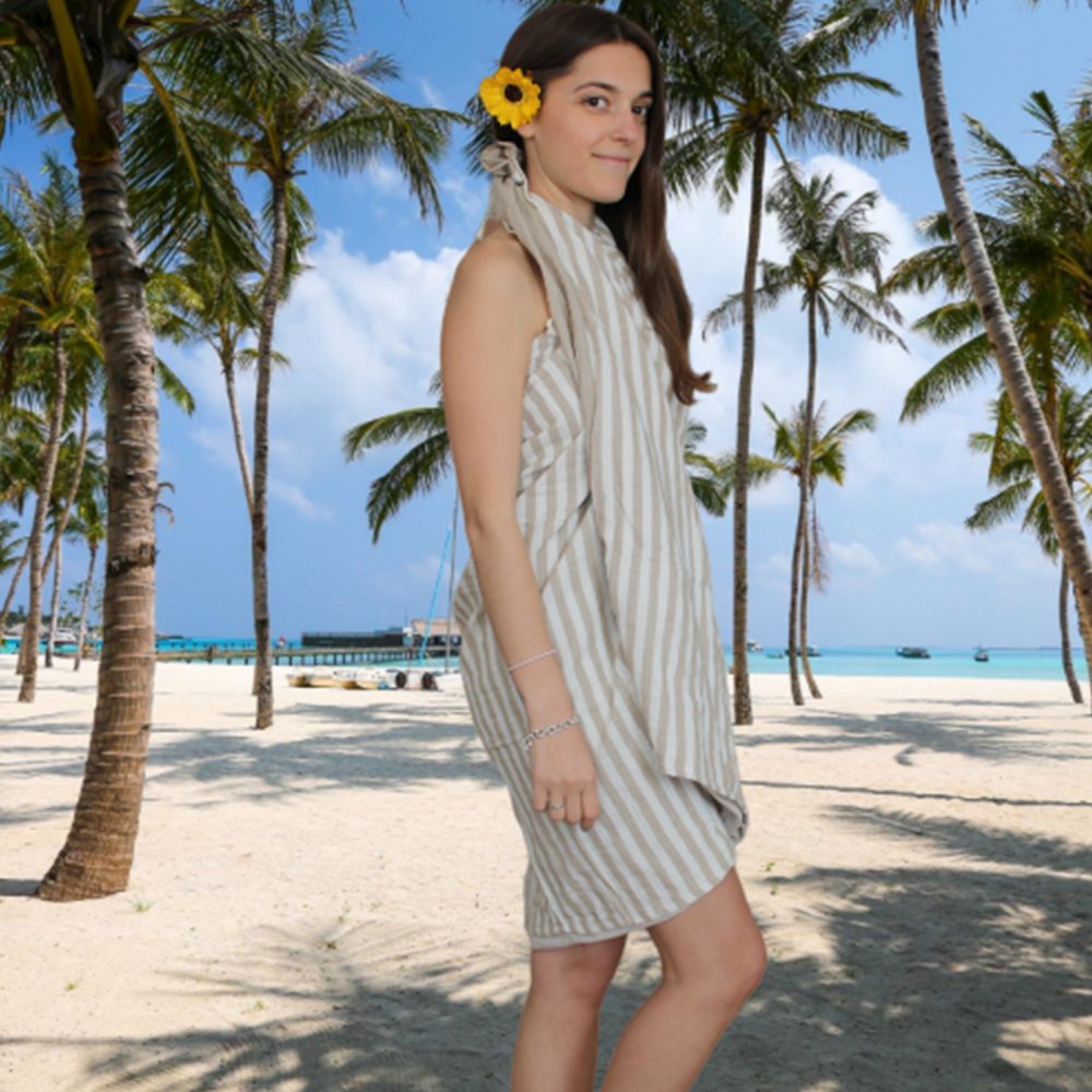 A woman models a beige and white striped towel dress on a tropical beach.