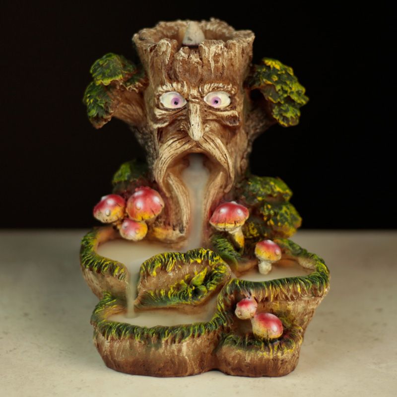 A tree-shaped incense holder with a carved face releases smoke. It resembles a tree trunk with moss, mushrooms, and greenery, set against a dark background on a light surface.