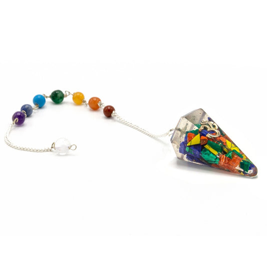 A chakra pendulum rests on a white surface. The pendulum features a cone-shaped crystal containing multicolored pieces, attached to a silver chain with seven colored beads, and a clear bead at the end. A silver “OM” symbol is on one facet of the crystal.