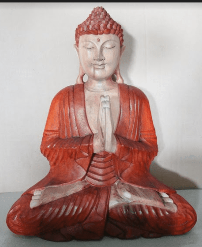 A red, intricately carved Buddha statue sits cross-legged with palms together in prayer, set against a plain, light-colored background.