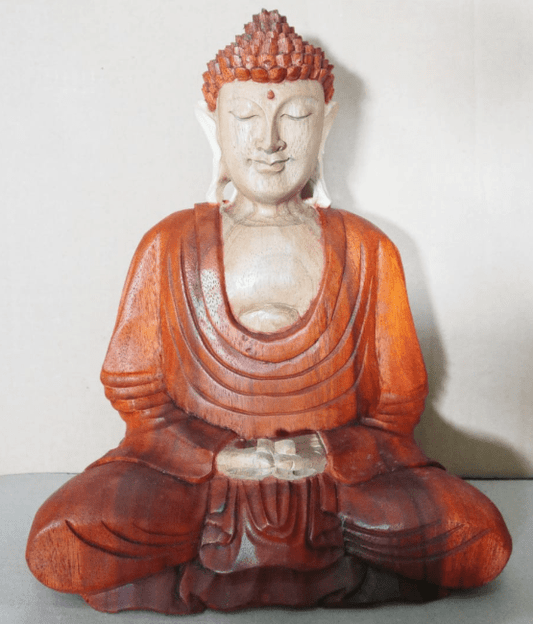 Wooden Buddha statue, sitting cross-legged with hands in a meditative posture, wearing a detailed robe. The background is a plain, light-colored surface.