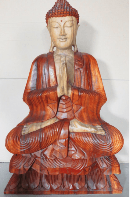 A carved wooden Buddha statue sits in a meditative pose with hands in prayer position, eyes closed, set against a plain background, conveying serenity and calm.